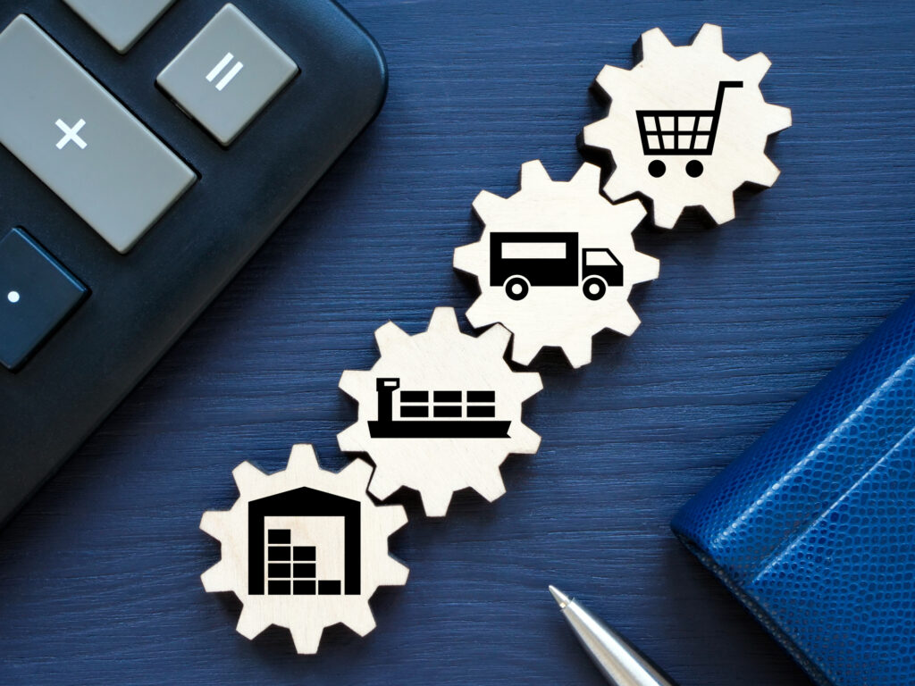 Four gears are laid out with images of a warehouse, a cargo ship, a transport truck and a shopping cart between a calculator and a pen and notebook as part of a product supply chain concept.